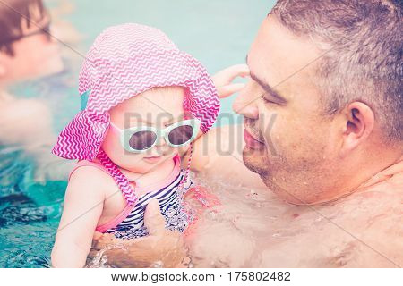 Family with cute baby girl faving fun in outdoor swimming pool on hot summer day.