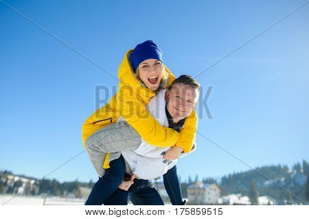 Young man holding his girlfriend on his shoulders. Girl embraces guy for neck. Young people look in the camera and cheerfully smile. Against the background of the blue sky.