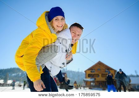 Young man holding his girlfriend on his shoulders. Girl embraces guy for neck. Young people look in the camera and cheerfully smile. Against the background of mountains and blue sky.