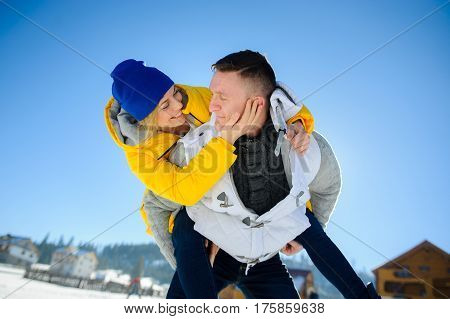 Young man holding his girlfriend on his shoulders. Girl embraces guy for neck. Young people look at each other with love and tenderness. Against the background of the blue sky.
