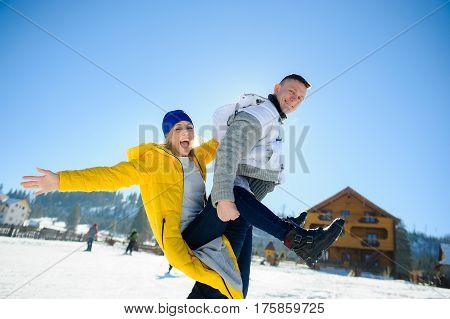 Guy and a girl are having fun in the courtyard of a cottage in a ski resort. A young man carries a girl on her back. She is laughing. Good winter day. Sun sky mountains.