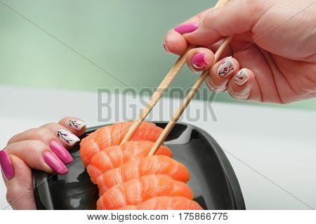 The female hand with magnificent manicure takes sushi with a light-salted salmon from a black ceramic plate by means of wooden sticks
