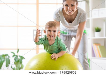 Happy mother and baby boy on fitness ball in nursery at home. Gimnastics for kids on fitball.