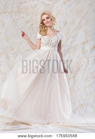 Portrait of the gorgeous smiling blonde bride with beautiful curly hair in a white lacy wedding dress on a light background. beautiful young model with bridal hairstyle and makeup throws up a white dress loop.ridal hairstyle and makeup in a white magnific