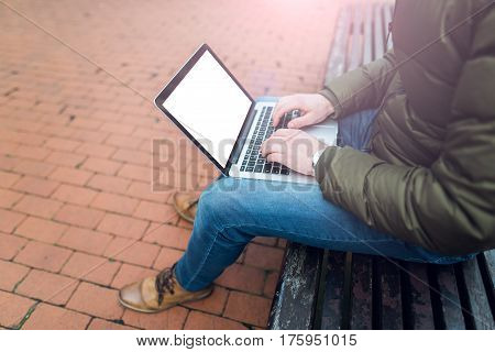 Laptop in a man's hands sitting on a wooden chair in park. Dressed in blue jeans and tan shoes. Mockup image of laptop with blank white screen. Photo with lens flare filter effect..