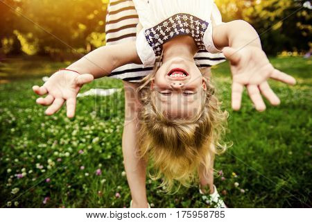 Young mom and cheerful adorable blond tot girl playing, having fun together in park in summertime, mother playfully holding her cute little daughter upside down, close up