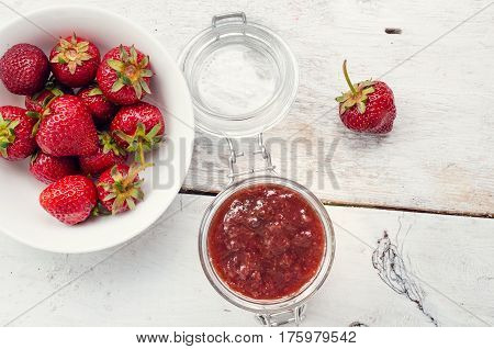 Homemade delicious strawberry jam in a glass jar and fresh strawberries on white rusric wooden background. Top view.