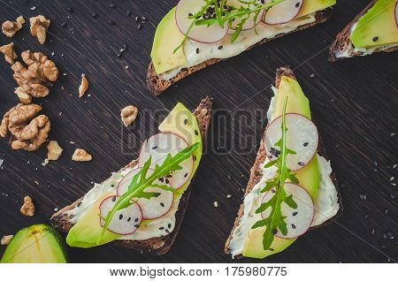 Sandwiches with avocado radish arugula cheese and nuts on a rye bread with sunflower seeds for healthy breakfast on dark wooden background. Vegetarian food. Top view.
