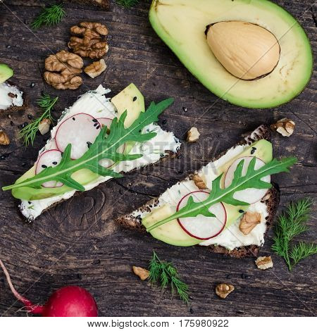 Sandwiches with avocado radish arugula cheese and nuts on a rye bread with sunflower seeds on wooden board on dark background. Healthy breakfast. Vegetarian food. Top view. Square.
