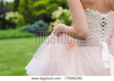 Close-up of a bride in a pink dress from the back. Woman stands with her back to the camera. Grass, trees and bushes in the background.