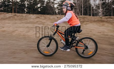 One Caucasian Children Rides Bike Road Track In Dirt Park. Girl Riding Black Orange Cycle In Racetra