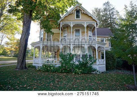 An Elegant Yellow Victorian Summer Cottage, With A Balcony Over Its Wraparound Porch, And A Corner T