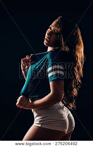 Sideways Portrait Of Beautiful Brunette Woman In Short Shorts And Blue T-shirt On Black Background.
