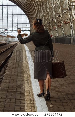 A Girl With A Raised Hand And A Suitcase In Retro Clothes Walks Along The Peron Station