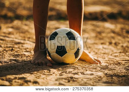 Close Up Picture Of An Old Ball And Foot Of A Poor Kid Who Is Playing Football In The Sunshine Day.