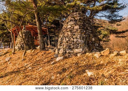 Two Stone Pyramids On Small Hill Under Grove Of Evergreen Trees And Wooden Gazebo In Background Loca