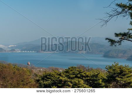 Landscape Of Reservoir On Hazy Overcast Day With Mountains, City Buildings And White Bridge In Backg