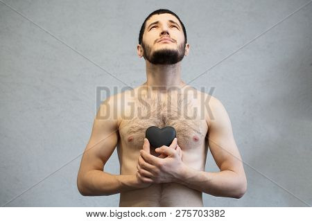 Naked Young, Bearded Man, Looking Up And Holding Black Heart In Hands Near Torso.