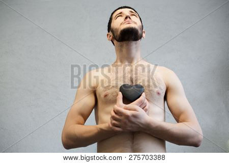Naked Young, Bearded Man, Looking Up And Holding Black Heart In Hands Near Torso.