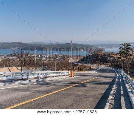 Landscape Of Country Road Leading To Reservoir On Hazy Overcast Day With Mountains, City Buildings A