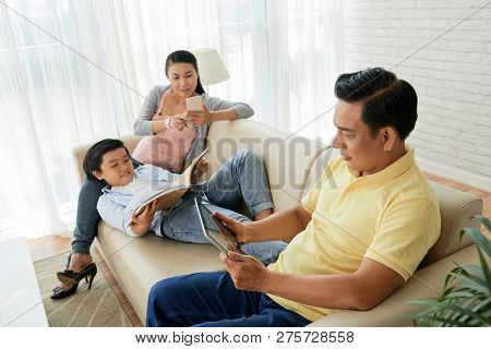 Teenge Boy Reading Book When His Parents Checking News Or Watching Videos On Their Gadgets
