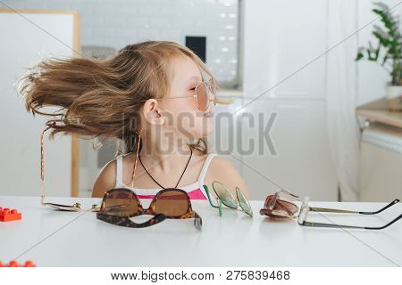 Portrait Of Cute Dark Blonde Little Girl With Short Hair Trying To Wear Pinhole Glasses. She Is At H