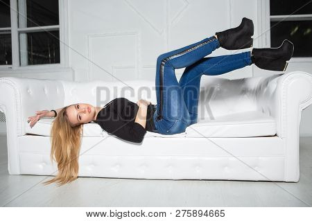 Cute Young Woman Posing Lying On The Sofa In The Studio, Dressed In A Black  T-shit And Jeans
