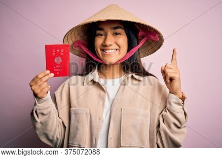 Young thai woman wearing traditional conical asian hat holding japan japanese passport surprised with an idea or question pointing finger with happy face, number one