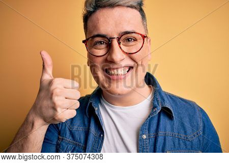 Close up of young handsome modern man wearing glasses and denim jacket over yellow background happy with big smile doing ok sign, thumb up with fingers, excellent sign