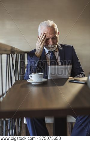 Senior Businessman Sitting At A Restaurant Table, Drinking Coffee And Reading Business News Using A 