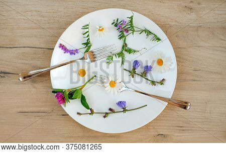 Grass And Flowers With Fork And Spoon In A White Plate On A Wooden Table, Vegetarian Food Concept.