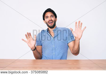 Handsome hispanic man wearing casual clothes sitting on the table afraid and terrified with fear expression stop gesture with hands, shouting in shock. panic concept. 