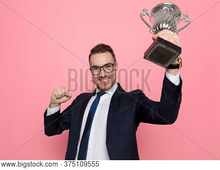 proud elegant businessman in suit wearing glasses holding up trophy and celebrating victory, holding fists in the air and standing on pink background