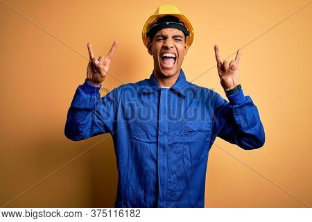 Young handsome african american worker man wearing blue uniform and security helmet shouting with crazy expression doing rock symbol with hands up. Music star. Heavy music concept.