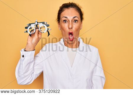 Middle age senior oculist woman holding optometrical glasses for vision correction scared in shock with a surprise face, afraid and excited with fear expression