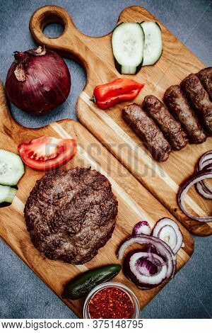Top View Of Wooden Kitchen Board With Grilled Meat, Turkish Cevap And Burger And Sliced Cucumber, To