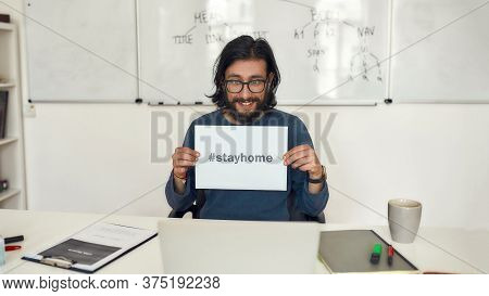 Young Happy Male Teacher Showing Paper With Text Stay Home At Webcam While Sitting At His Workplace 