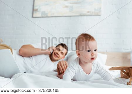 Selective Focus Of Man Talking On Smartphone While Touching Cute Little Son Crawling On Bed