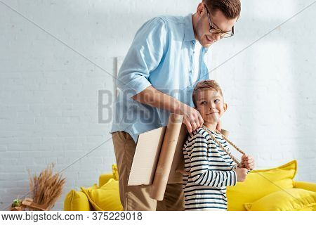 Father Fixing Carton Plane Wings On Back Of Adorable Son