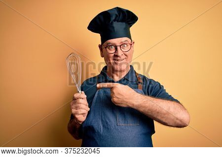 Middle age hoary cooker man wearing apron and hat holding whisk over yellow background very happy pointing with hand and finger