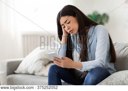 Waiting For His Call. Upset Asian Girl Looking Sadly At Mobile Phone Screen, Sitting On Sofa At Home