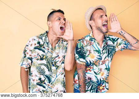 Young gay couple of two men wearing summer hat and hawaiian shirt shouting and screaming loud to side with hand on mouth. communication concept. 