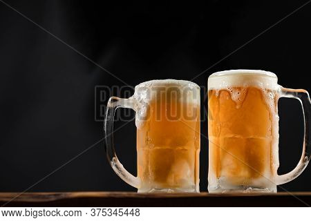Two Cold Mugs With Beer, With Overflowing Foam, On Wooden Table And Dark Background, Space For Writi