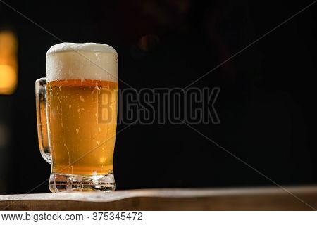 Cold Mug With Beer, With Overflowing Foam, On Wooden Table And Dark Background