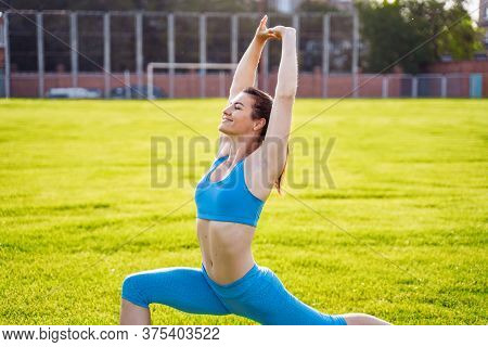 Attractive, Athletic Girl In A Blue Suit Stretches In The Stadium From Green Grass. Enthusiastically
