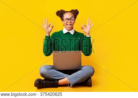 Im Fine. Excited Afro Teenage Girl Showing Ok Sign, Sitting On Floor With Laptop Over Yellow Backgro