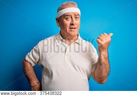 Middle age hoary sportsman doing sport wearing sportswear over isolated blue background smiling with happy face looking and pointing to the side with thumb up.
