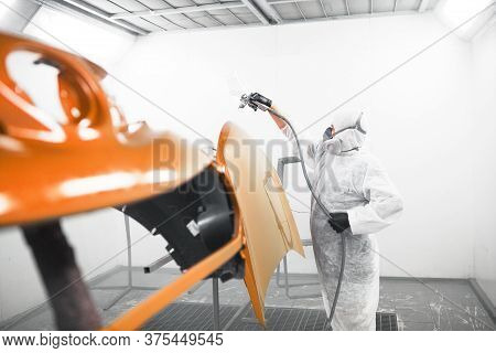 Man In Protective Mask And Clothes Sprays Varnish To Car Hood With A Spray Gun In A Paint Booth.
