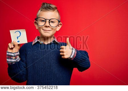 Young little caucasian kid wearing glasses holding paper note with question mark sign happy with big smile doing ok sign, thumb up with fingers, excellent sign