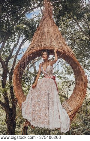 Beautiful Bride In Her Wedding Dress Posing For Photos On A Wooden Swing In The Tree.